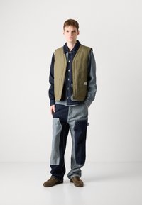 Green padded vest over a denim shirt, paired with patchwork denim pants in blue tones. Brown shoes complete the outfit.