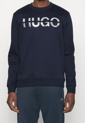 Mand iført en mørkeblå sweatshirt med skinnende sølv "HUGO"-logo på brystet og matchende mørkeblå bukser.