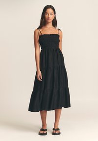 Abito lungo nero a balze con corpetto arricciato, spalline sottili e tessuto strutturato. Indossato con sandali neri.