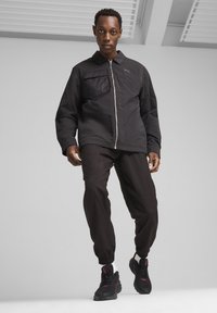 Sort zip-up jakke med en brystlomme, parret med løstsiddende sorte bukser og sorte sneakers med røde accenter. Glat stofstruktur.