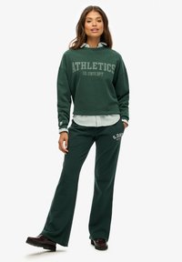 Sudadera verde con el texto "ATHLETICS" y una camisa de botones a rayas debajo, combinada con pantalones anchos verdes a juego y zapatos oscuros.