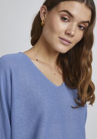 Pull en tricot bleu clair avec un col en V et de longues manches amples. Texture douce et motif de point subtil. Boucles d'oreilles en or viennent compléter le look.