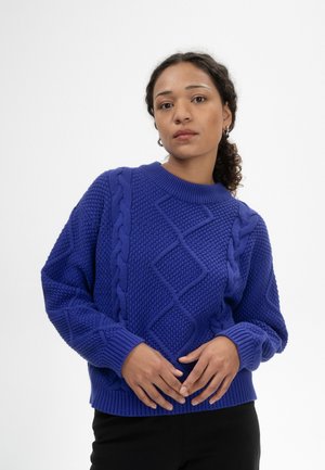 Femme aux cheveux bouclés portant un pull en maille bleue texturée avec des motifs en losanges et en torsades, debout devant un fond blanc uni.