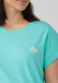 Turquoise t-shirt met korte mouwen en een ronde hals, met een kleine witte visafbeelding op de linkerkant van de borst. Zacht, lichtgewicht stof met een gestructureerde afwerking.
