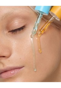 Serumpipetten in Blau und Gelb geben klare und gelbe Öle auf glatte, helle Haut, während glitzernde Tropfen das Gesicht hinunterlaufen.