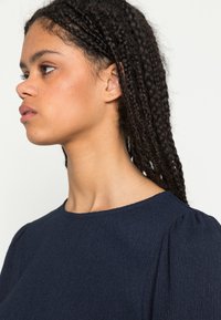 Top azul marinho com um acabamento texturizado, decote redondo e mangas ligeiramente bufantes. O material parece leve e ligeiramente amarrotado.