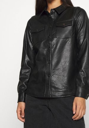 Femme portant une chemise boutonnée en similicuir noir avec deux poches poitrine, associée à un pantalon foncé, debout devant un fond clair.