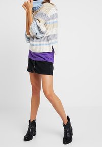 Kvinna i en randig stickad tröja, lila topp, svart minikjol och svarta ankellånga stövlar som står mot en neutral bakgrund.