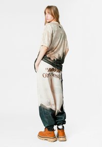 Camiseta gráfica de gran tamaño en beige y negro con tejido texturizado; combinada con pantalones beige de corte holgado que presentan acentos oscuros y texto audaz. Botas marrones.