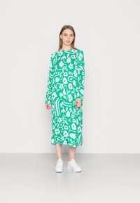 Vestido verde de manga longa com padrões florais brancos e abstratos, apresentando uma silhueta reta e combinado com ténis brancos.