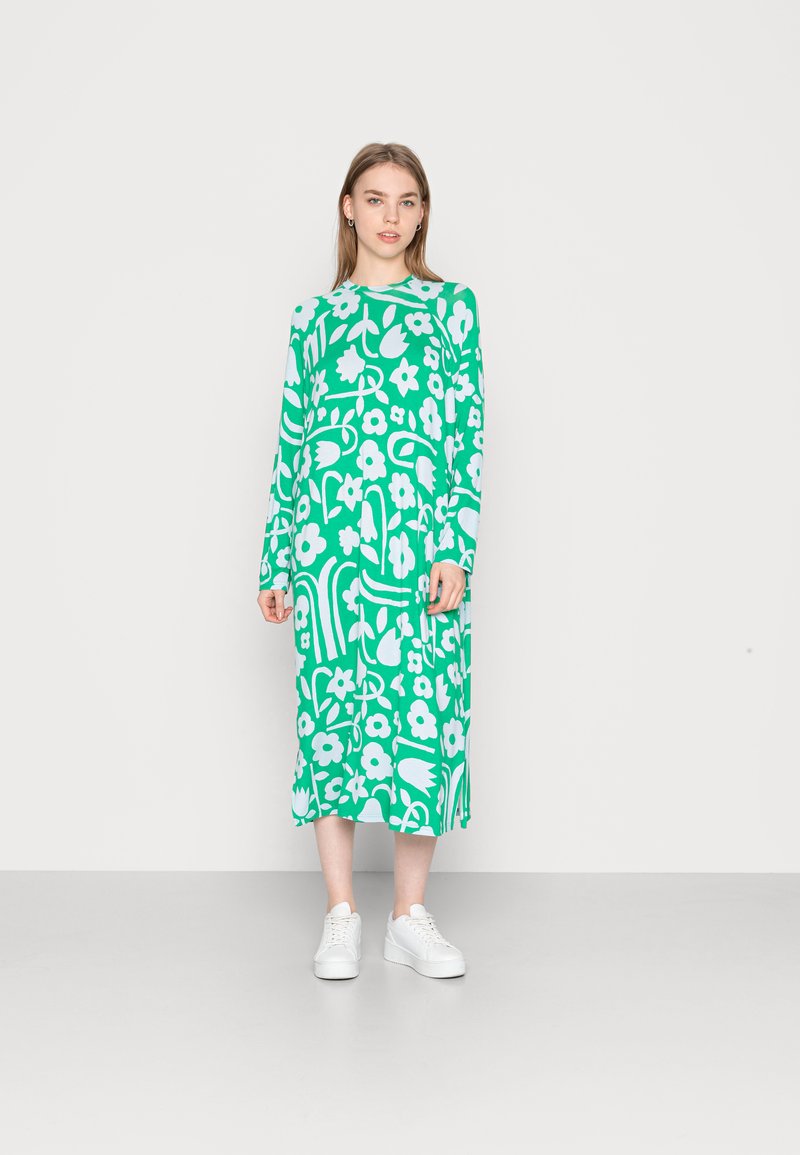 Vestido verde de manga longa com padrões florais brancos e abstratos, apresentando uma silhueta reta e combinado com ténis brancos.