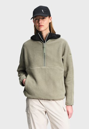 Woman wearing light olive fleece pullover with front zipper, black hood, black cap, and light beige cargo pants, standing with one hand in pocket.