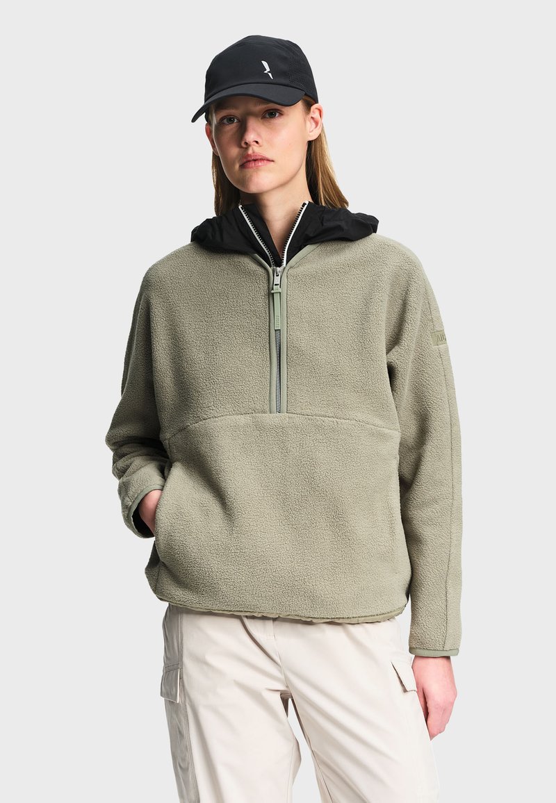 Woman wearing light olive fleece pullover with front zipper, black hood, black cap, and light beige cargo pants, standing with one hand in pocket.