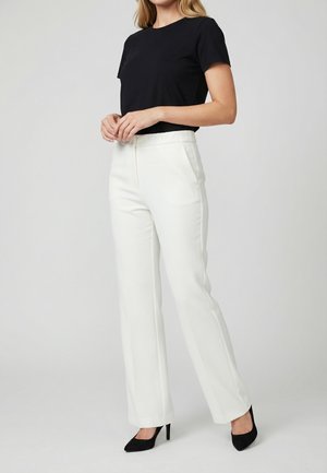 Femme portant un haut noir à manches courtes, un pantalon blanc droit et des talons hauts noirs pointus, debout devant un fond uni.