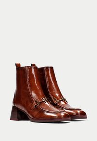 Botas de tobillo de cuero marrón con un acabado brillante, que cuentan con un tacón bloque y acentos decorativos de hardware dorado.