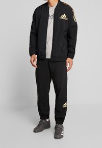 Svart bomberjacka med gulddetaljer och logotyp, ihop med svarta joggers med guldinslag, kompletterat av mörka träningsskor.