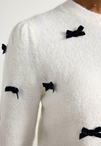 Pullover bianco a maglia con finitura testurizzata, caratterizzato da piccoli fiocchi in velluto blu navy disposti decorativamente sulla parte anteriore.
