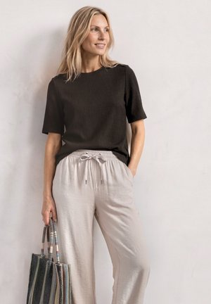 Femme aux cheveux blonds portant un haut noir à manches courtes et un pantalon beige clair à cordon, tenant un sac fourre-tout tissé rayé à ses côtés.