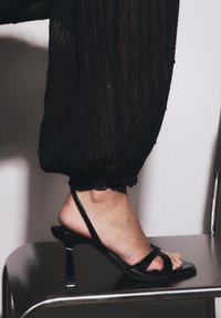 Schwarze Riemchensandale mit dünnem Absatz und Knöchelriemen. Glattes Lackmaterial, offene Zehenpartie, gezeigt an einem Fuß auf einem metallischen Stuhl.