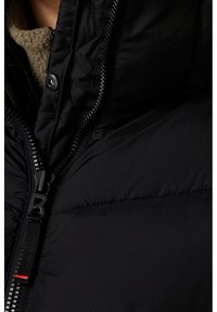 Chaleco puffer negro con un alto cuello, cierre de cremallera con un tirador y una textura lisa y brillante. Detalle de costura notable.
