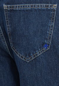 Πίσω τσέπη τζιν σε σκούρο μπλε, με ανοιχτό καφέ ραφή και ένα μικρό μπλε κεντητό λογότυπο στη γωνία.