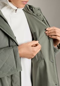 Khaki prešita jakna z detajli z zadrgo, mehak material, gumbki na pritisk, sproščen kroj, nosi se čez belo majico.