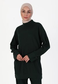 Maglione a coste verde scuro con maniche lunghe e collo alto; presenta una texture verticale e spacchi laterali all'orlo.