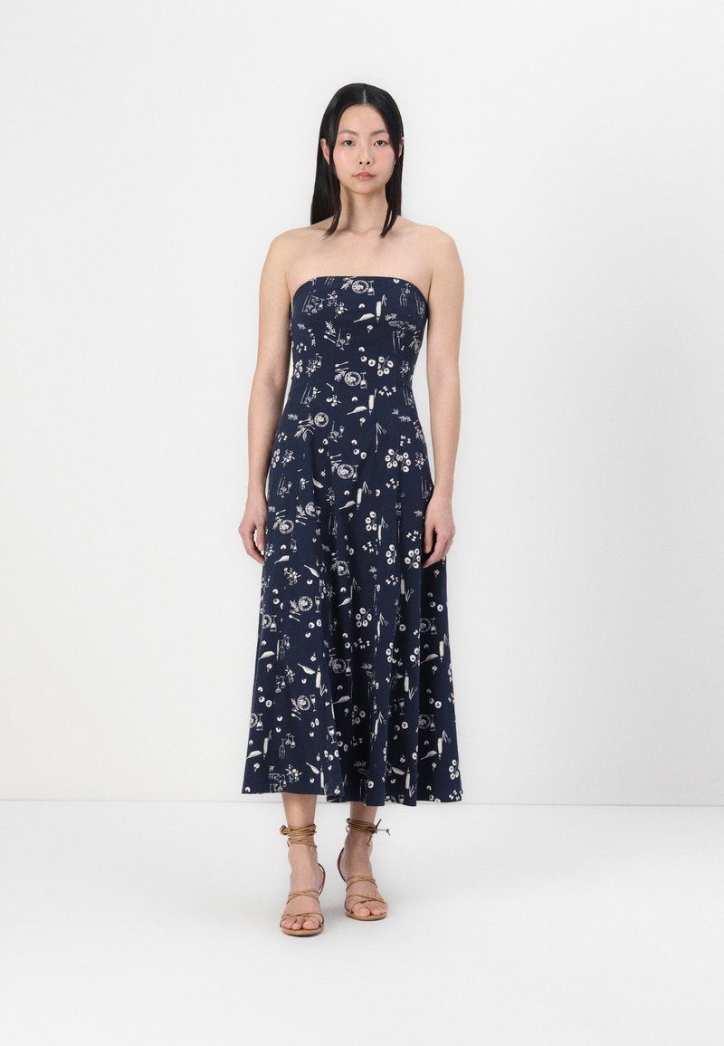Mörkblå strapless maxiklänning med blommönster; figursydd korsett, utställd kjol, lätt tyg. Modellen bär sandaler med ankelrem i beige.
