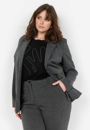 Šedý oblekový sako s odpovídajícími kalhotami, vyrobené z jemného materiálu. Černá košile pod ním má design se stříbrnými pruhy.