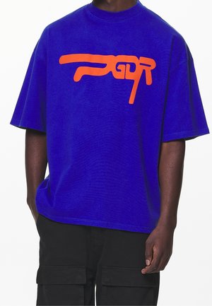 Person iført en lyseblå oversized T-shirt med et abstrakt orange mønster på brystet og sorte cargobukser med lommer.