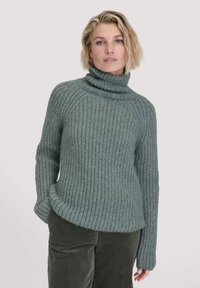 Hellgrüner, gestrickter Pullover mit hohem Rollkragen und geripptem Muster, mit lockerem Schnitt und leicht abgerundetem Saum, kombiniert mit dunklen Cordhosen.