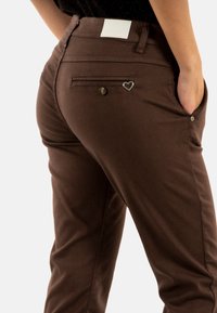Pantaloni di cotone marroni con vestibilità slim, dotati di una tasca posteriore, accessori in metallo a forma di cuore e un'etichetta bianca sulla cintura.