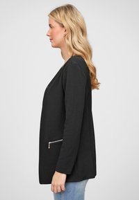 Svart strukturert blazer med glatt front, åpen design, lange ermer og sidelomme med glidelås, vist fra en sidevinkel mot en lys bakgrunn.