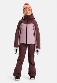 Snow jacket in purple and burgundy, paired with matching pants. The wearer holds gloves with a colorful pattern and wears an orange beanie.