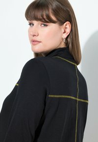 Vrouw met lichtbruin haar en zilveren oorbellen in de vorm van een ring, gekleed in een zwarte top met gele gestikte kruisdetails op de achterkant, terwijl ze over haar schouder kijkt.