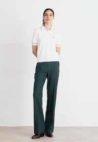 Witte polo met gele accenten en korte mouwen, gecombineerd met donkergroene, op maat gemaakte broeken. Beide zijn gemaakt van een gladde stof.