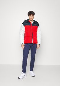 Chaqueta acolchada roja y blanca con capucha azul oscura, cierre frontal y logo en el pecho, combinada con pantalones azul marino y zapatillas blancas.