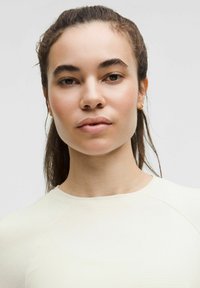 Mudeli pikk pruun juustega, seljas heledast kreemjast tekstuuriga särk ümarate kaelustega. Näha on kullast rõngassõrmuseid.