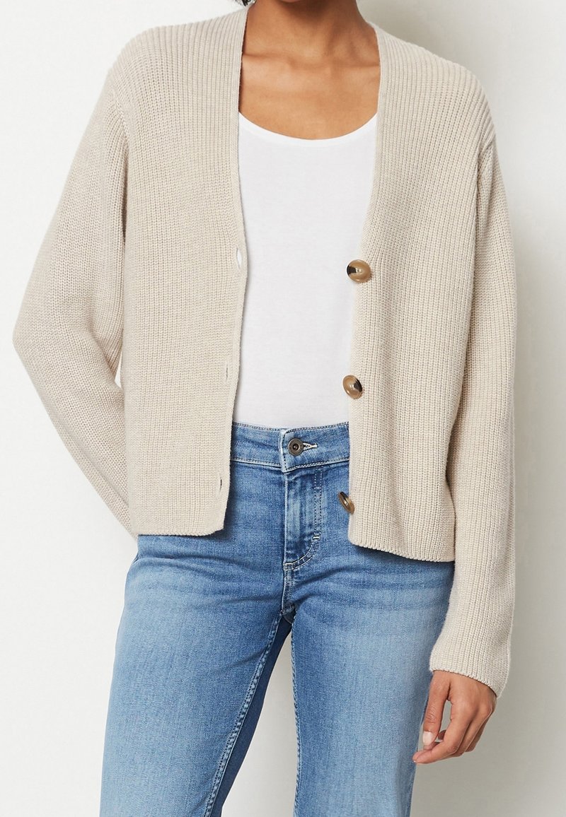 Beżowy dziergany kardigan z prążkowaną teksturą, z trzema dużymi brązowymi guzikami, otwartym przodem i długimi rękawami, noszony na białej bluzce.