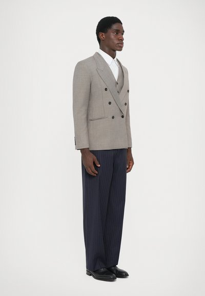 Grauer doppelt brustiger Blazer mit schwarzen Knöpfen, kombiniert mit navyblauen, gestreiften weit geschnittenen Hosen und schwarzen Schuhen, getragen über einem weißen Hemd.