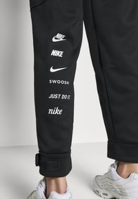Černé tepláky s několika bílými logy Nike, včetně "Swoosh" a "Just Do It," a detailem na suchý zip u kotníku.