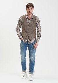 Gilet a scacchi marrone sopra una camicia a fantasia floreale, abbinato a jeans chiari strappati e sneakers bianche.