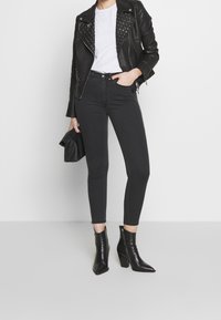 Chaqueta de motorista de cuero negra con tachuelas plateadas, camiseta blanca, jeans ajustados negros y botas de tobillo puntiagudas. Modelo sosteniendo un clutch negro.