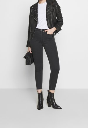 Chaqueta de motorista de cuero negra con tachuelas plateadas, camiseta blanca, jeans ajustados negros y botas de tobillo puntiagudas. Modelo sosteniendo un clutch negro.