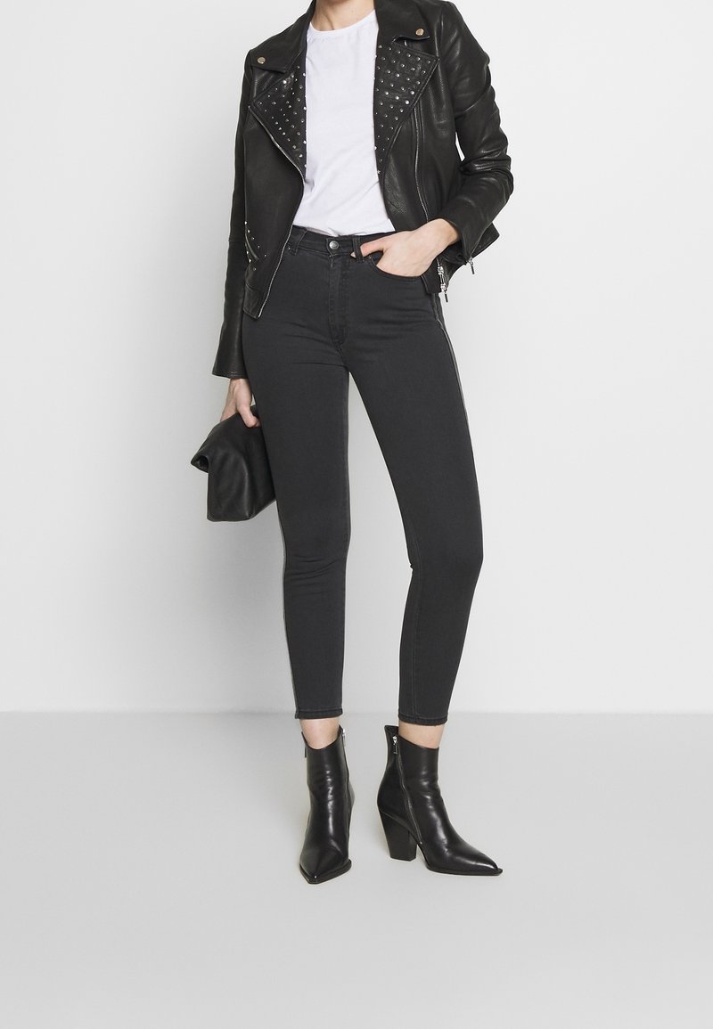 Chaqueta de motorista de cuero negra con tachuelas plateadas, camiseta blanca, jeans ajustados negros y botas de tobillo puntiagudas. Modelo sosteniendo un clutch negro.