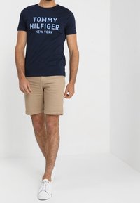 Navy blue cotton t-shirt with "TOMMY HILFIGER NEW YORK" printed in light blue; paired with beige shorts and white sneakers.