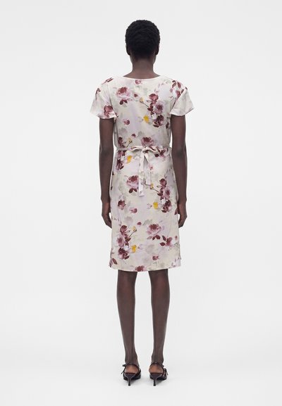 Γυναίκα με λευκό φόρεμα μέχρι τα γόνατα με ροζ και κίτρινo λουλουδάτο σχέδιο, κοντά μανίκια, δεμένη μέση και μαύρα σανδάλια με τακούνι, γυρισμένη από πίσω.