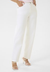 Jeans de denim blanco con corte recto, cinco bolsillos, cierre frontal con botón y detalle de costura sutil. Combinados con tacones ligeros y con tiras.