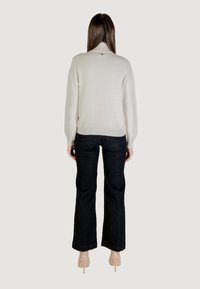 Pull en tricot crème avec un col montant, présentant un léger scintillement, associé à un pantalon en denim foncé évasé et des talons nude. Vue de dos.