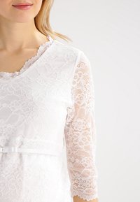 Chemisier en dentelle blanche avec des manches trois-quarts, un décolleté en feston et une ceinture décorative à la taille, orné de motifs floraux.